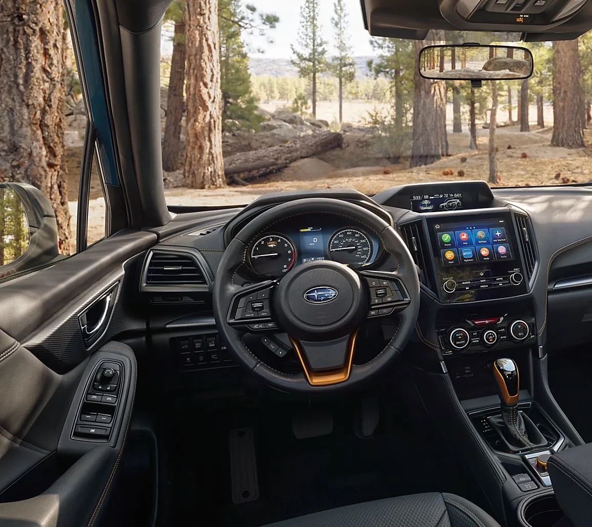 Habitacle du Subaru Forester Wilderness 2025 avec intérieur noir et surpiqûres orange, vue sur une forêt à travers les fenêtres.
