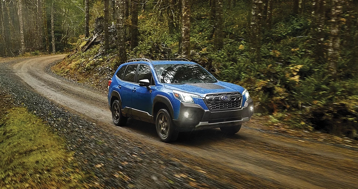 Subaru Forester Wilderness 2025 bleu roulant sur un chemin forestier sinueux, entouré d’arbres verdoyants et de brouillard léger.